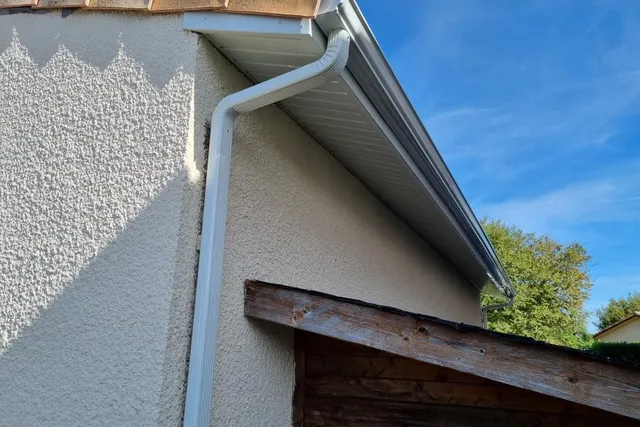 Façade en crépi clair sous une avancée de toit, avec sa gouttière et sa descente d'eau blanches
